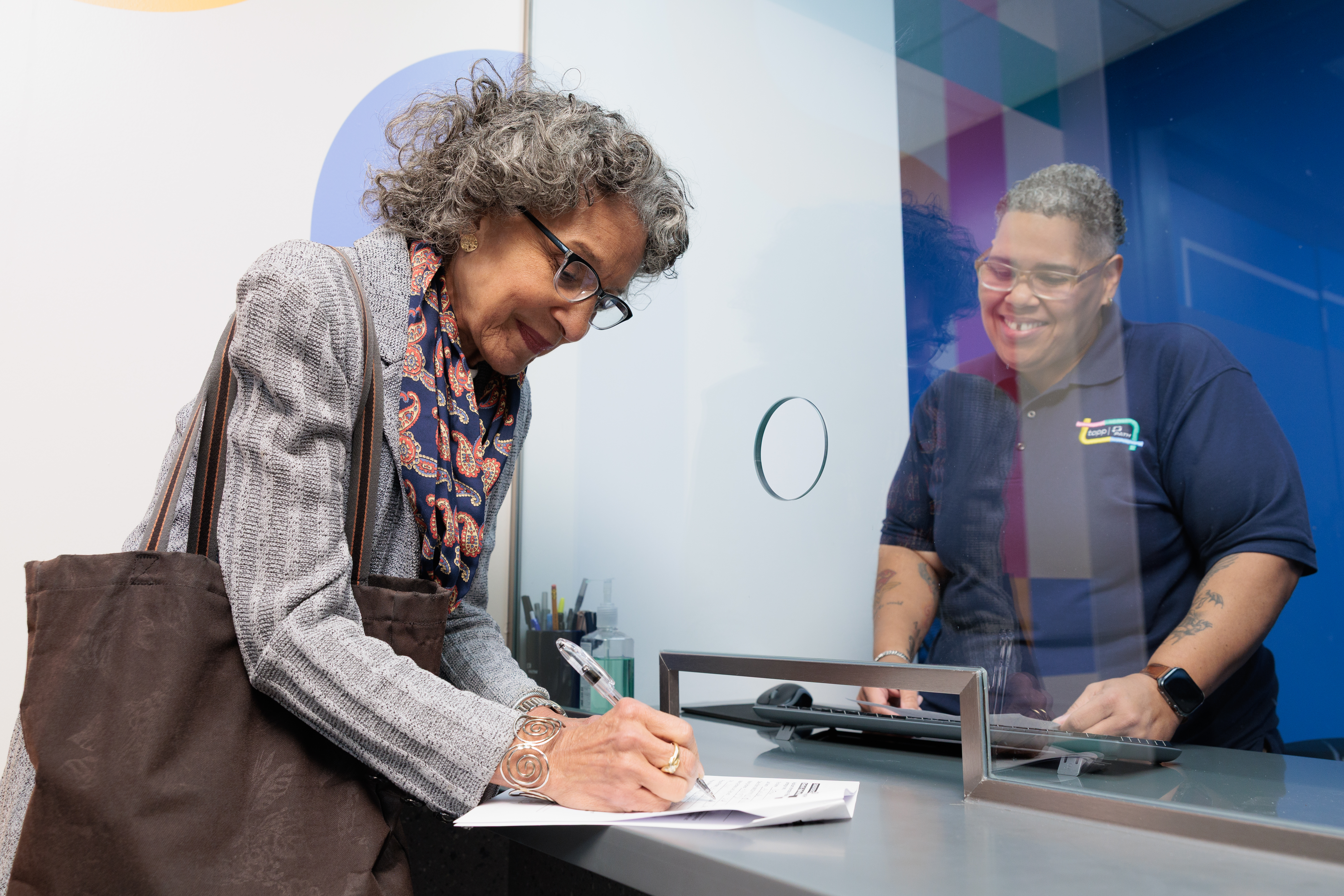 An older woman with curly gray hair, glasses, and a paisley scarf leans over a service counter, filling out a form with a pen. Behind the glass partition, a smiling PATH transit employee in a navy polo shirt assists her from the other side.
